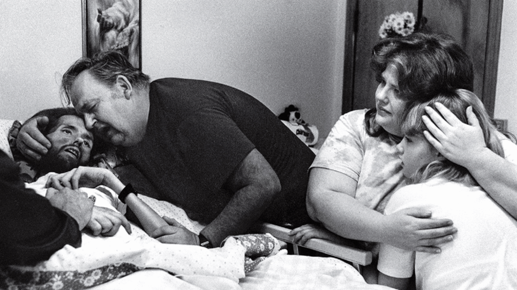 A man dying of AIDS, David Kirby, lies on his deathbed surrounded by family members; the scene powerfully expresses the tragedy of the disease and family unity.